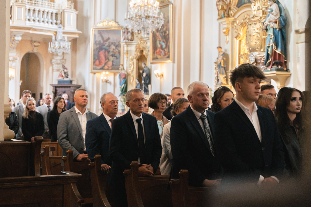 Goście stojący w ławkach podczas ślubu w ozdobnym wnętrzu kościoła.
